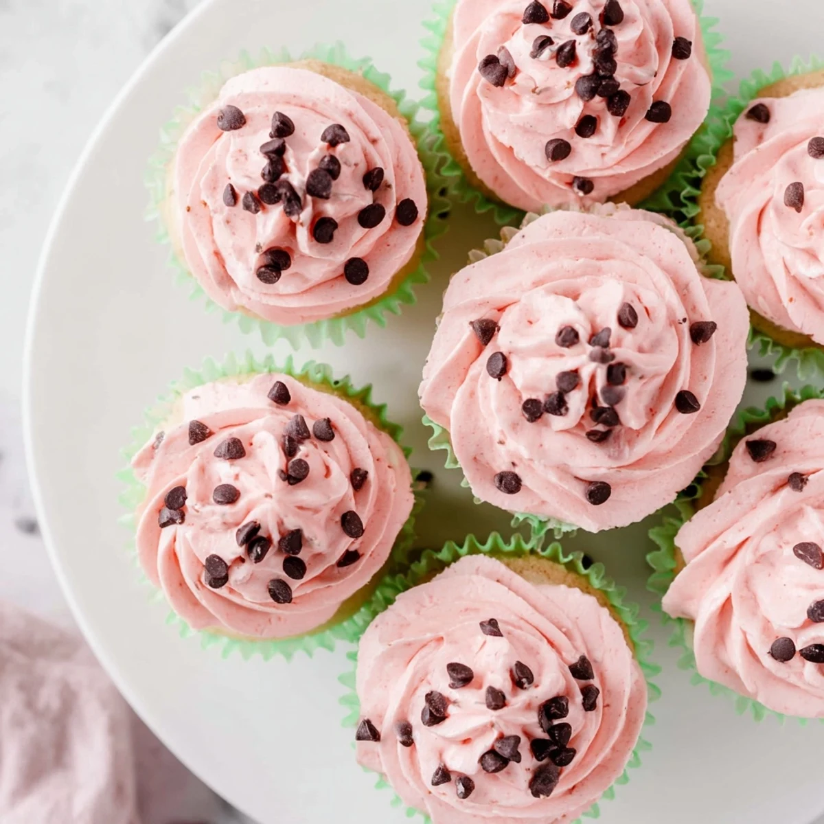 Pink frosted watermelon cupcakes topped with chocolate chip seeds on a white plate