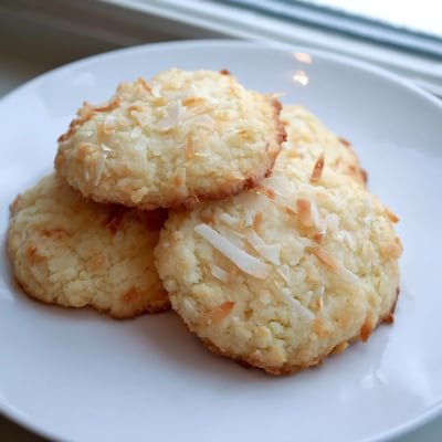 Chewy coconut cookies stacked on a plate dusted with powdered sugar crumbs.